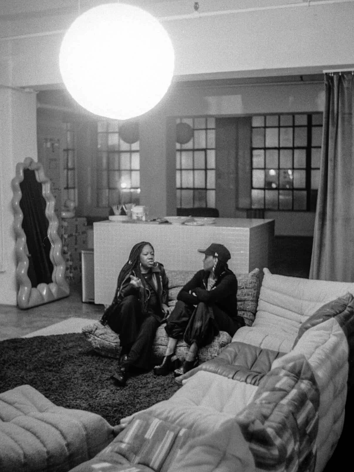 Two women sitting and talking on a couch in a living room, with large windows, curtains, a decorative mirror, and cozy furniture.