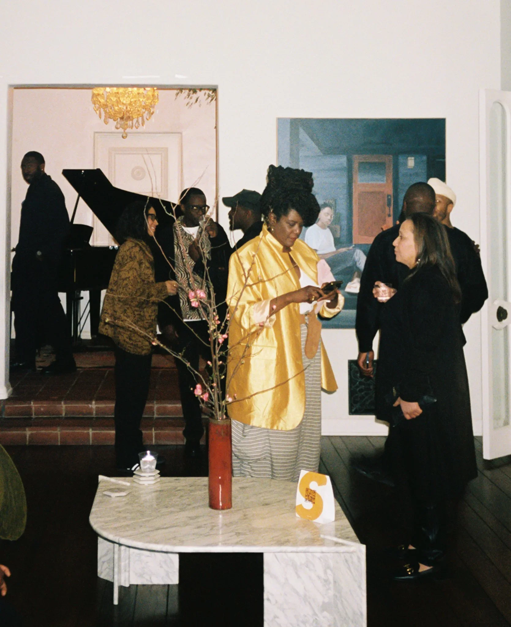 Group of people socializing around a table at an indoor event, with a woman in a yellow jacket looking at her phone and a man playing a piano in the background.