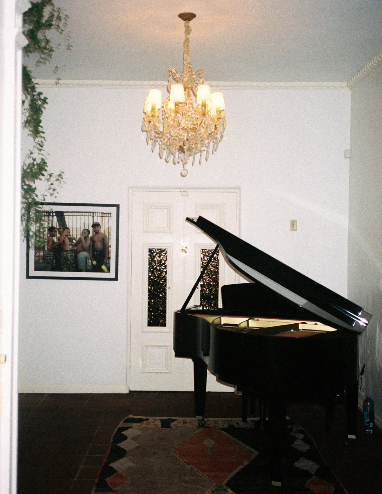 Interior of a home with a grand piano, chandelier, artwork, and a door at the back. The chandelier is ornate with multiple lamps, hanging from a decorative ceiling. A framed photograph of children is on the wall to the left. A colorful rug is on the floor in front of the piano.