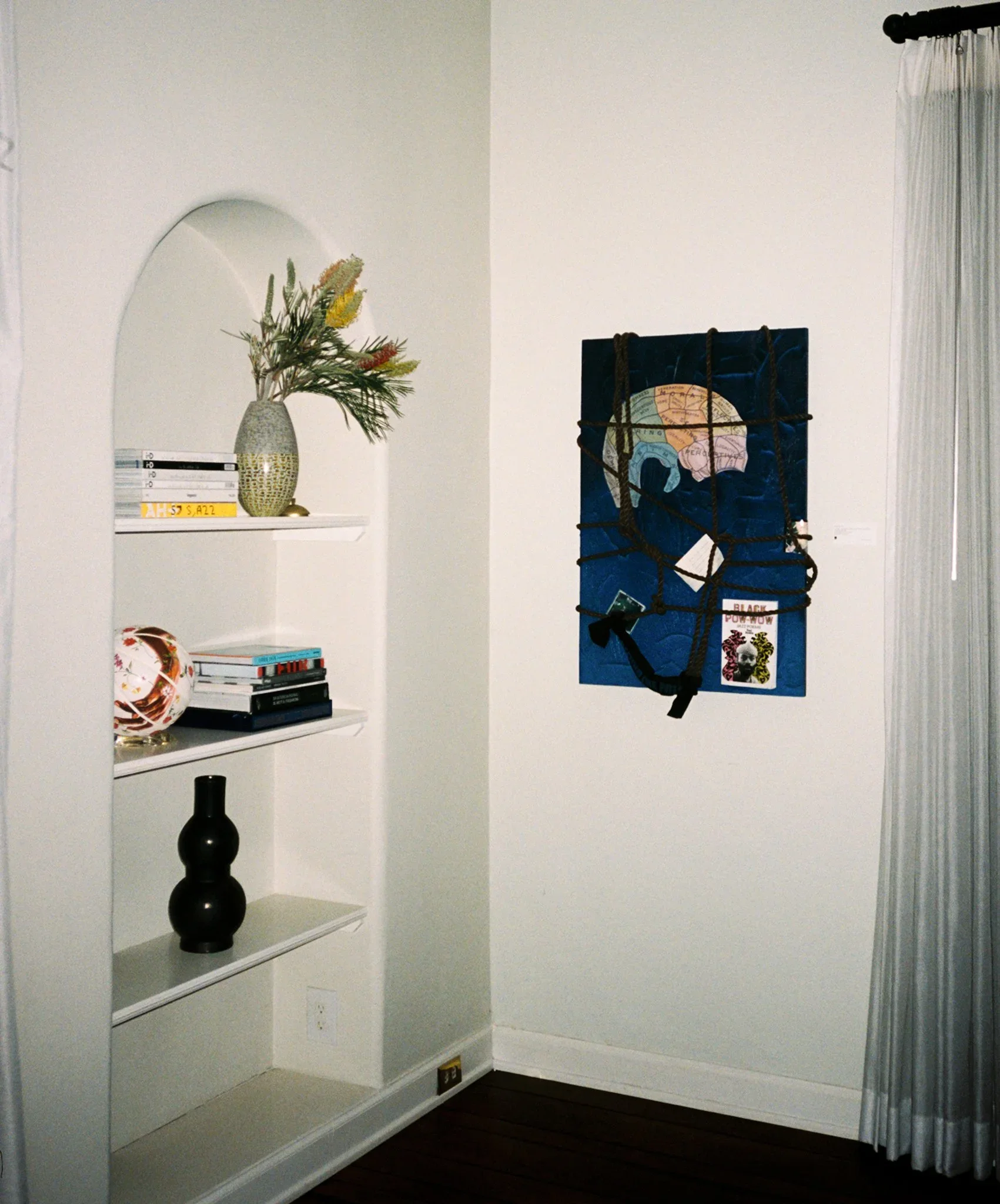 A white corner wall with three floating shelves, decorated with a vase of flowers, books, and a ceramic ornament, with a white curtain on a window and a black decorative vase on the floor.