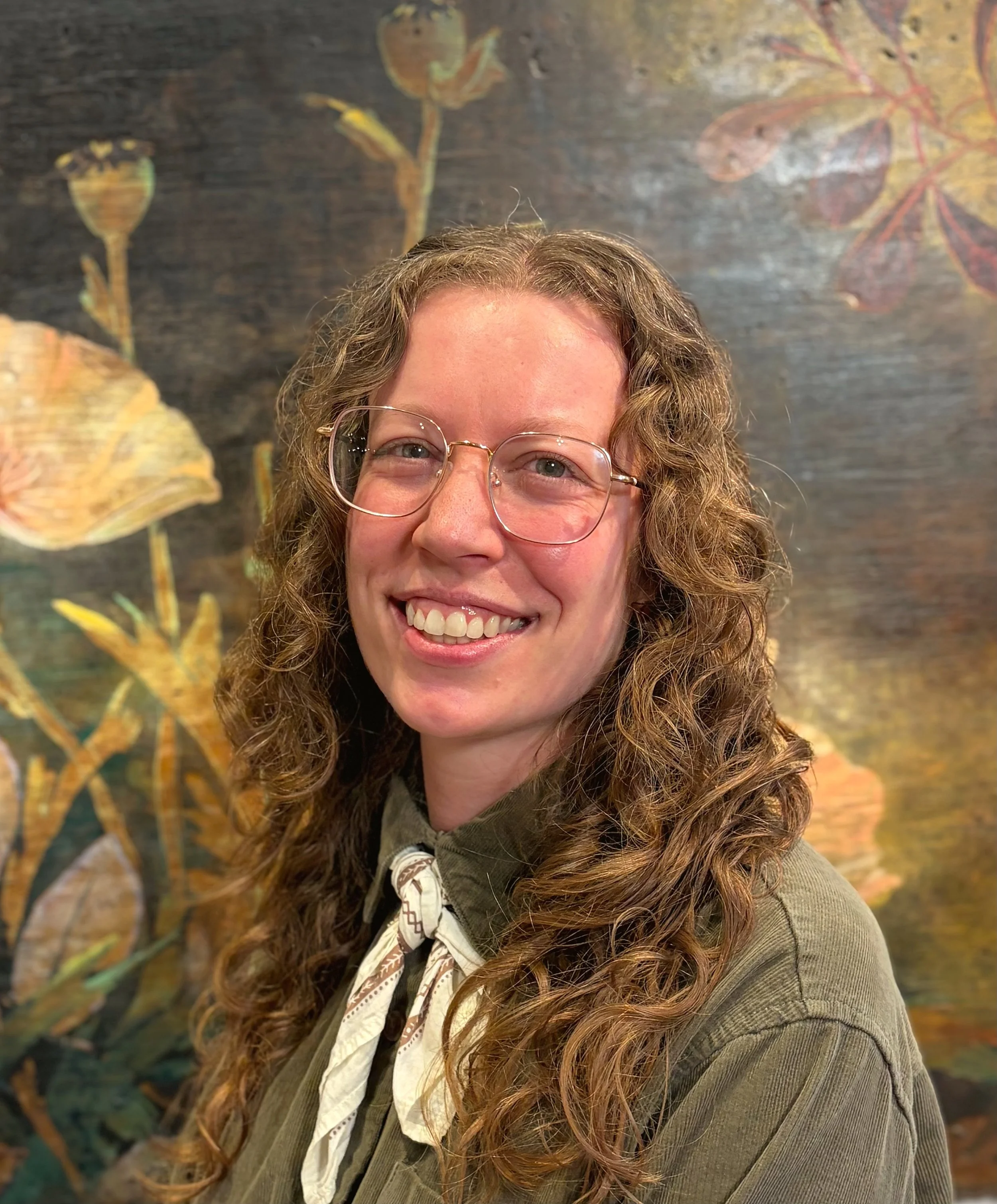 A blonde woman with curly hair and glasses smiles in front of a painting of flowers.