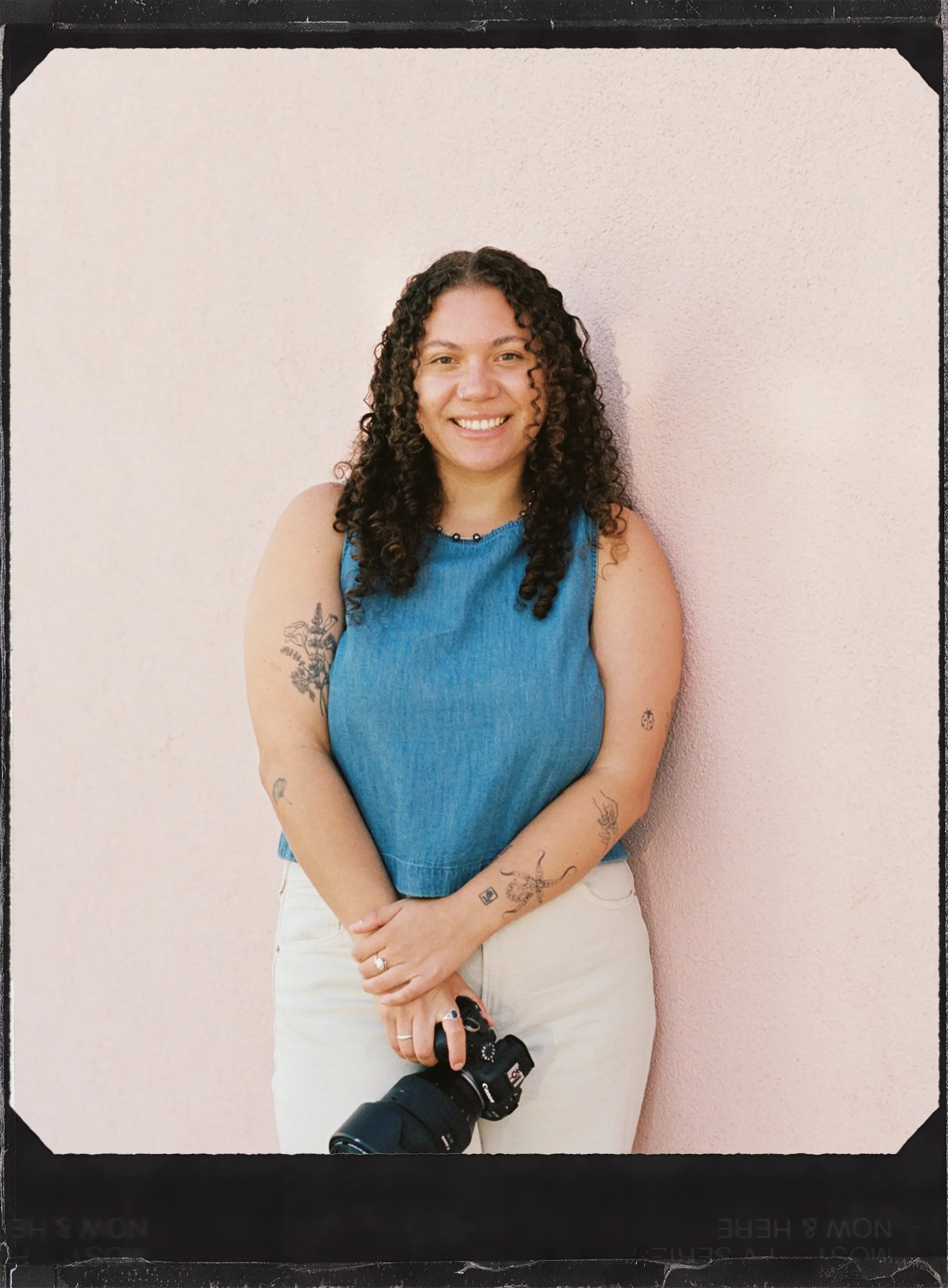 A woman with curly hair, wearing a blue sleeveless top and white pants, holding a camera and a camera strap, standing against a pink wall and smiling at the camera.