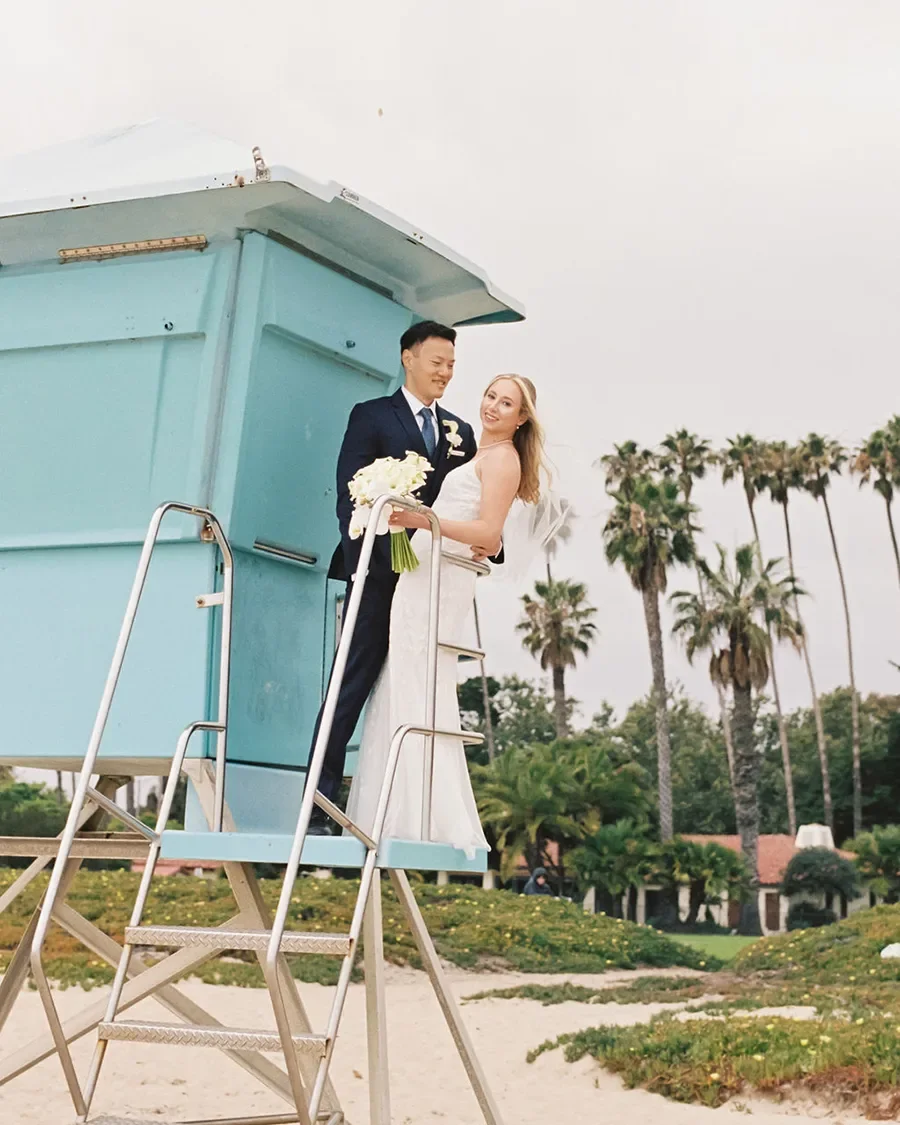 A newlywed couple standing on a lifeguard tower, holding a bouquet of white flowers, with palm trees in the background, on a beach or coastal area.
