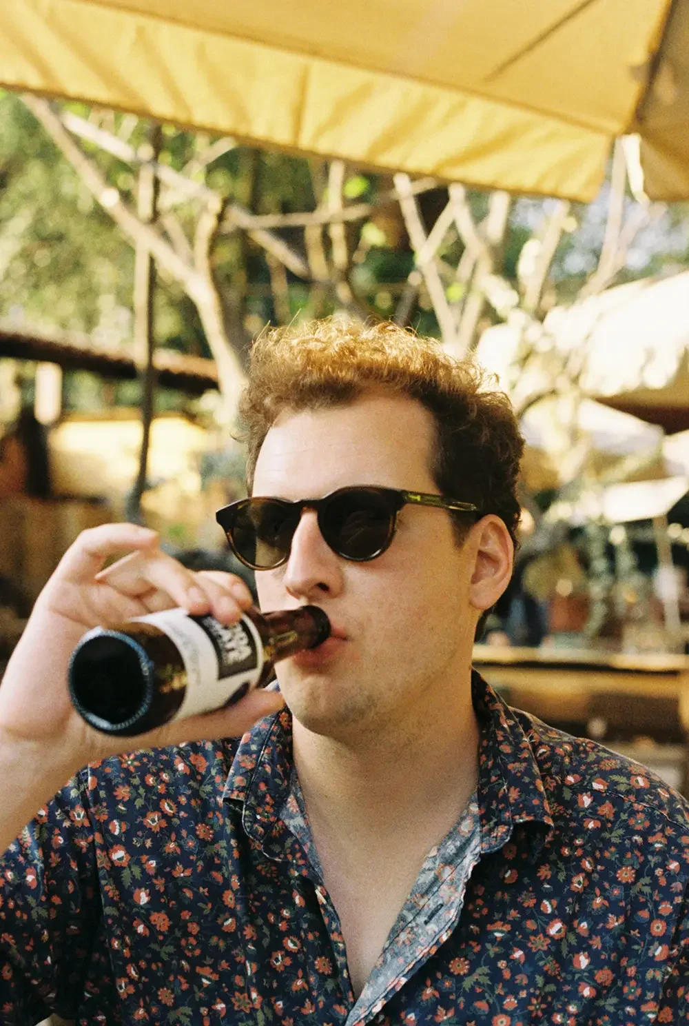 Zoe's partner, Patrick, wearing sunglasses and a floral shirt, drinking from a brown beer bottle at an outdoor setting with trees and umbrellas.