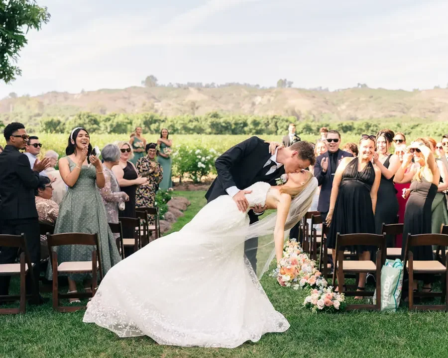A newlywed couple sharing a kiss outdoors at a wedding ceremony with guests clapping and smiling.