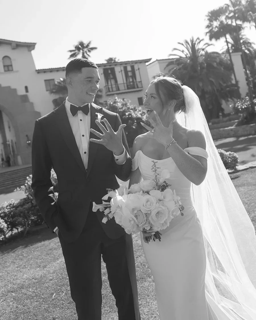 A bride and groom are smiling and showing their wedding rings outdoors on a sunny day. The bride holds a bouquet of white roses and greenery, and her veil flows down her back. Background includes palm trees and European-style buildings.