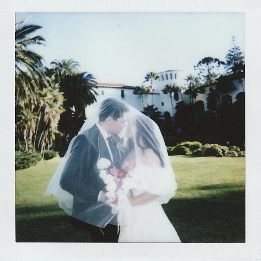 A bride and groom sharing a kiss outdoors on a sunny day, with palm trees and a white building in the background.
