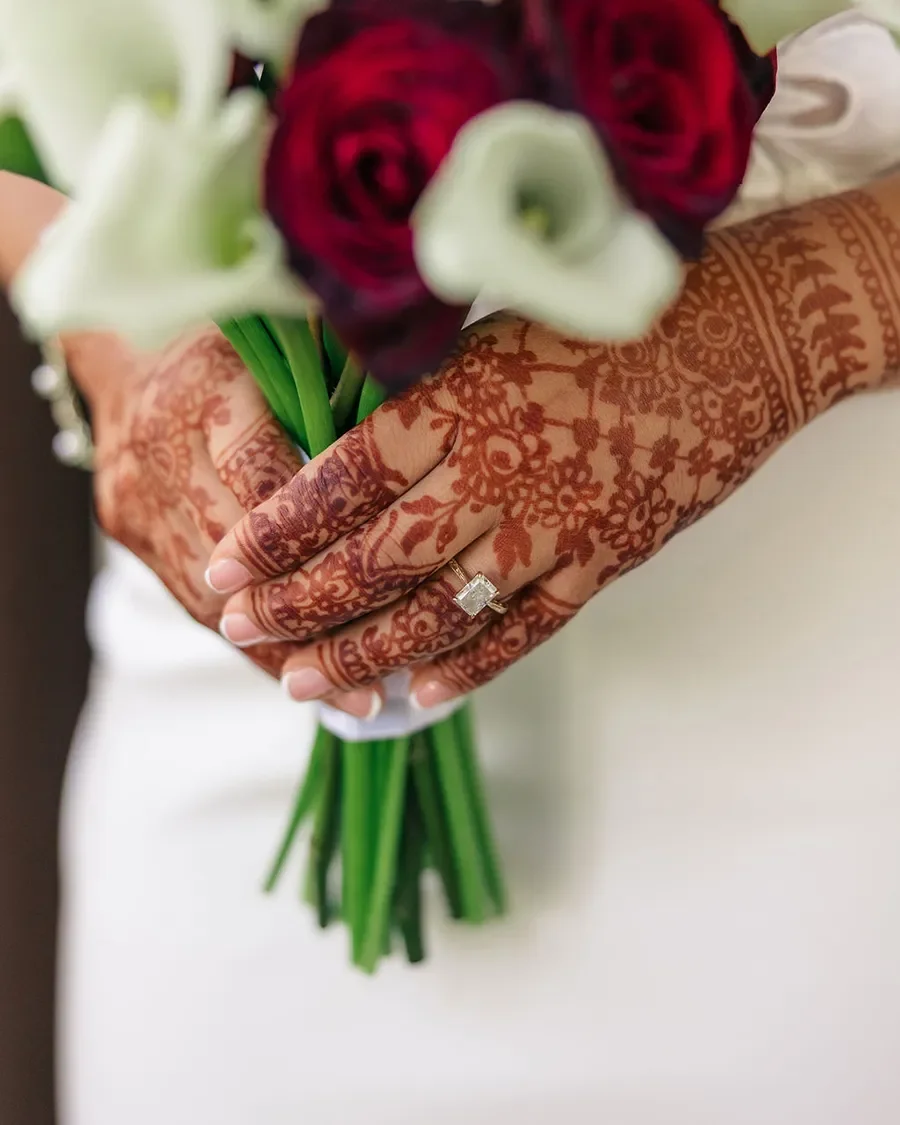Close-up of a person's hand with intricate henna designs holding a bouquet of red and white flowers.