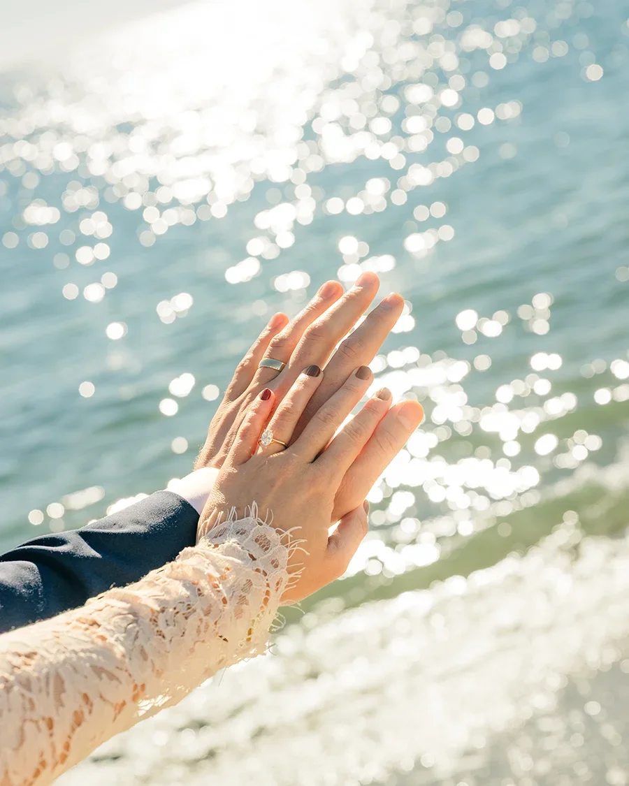 Close-up of two couples' hands with wedding rings, touching over the ocean with sunlight reflecting off the water.