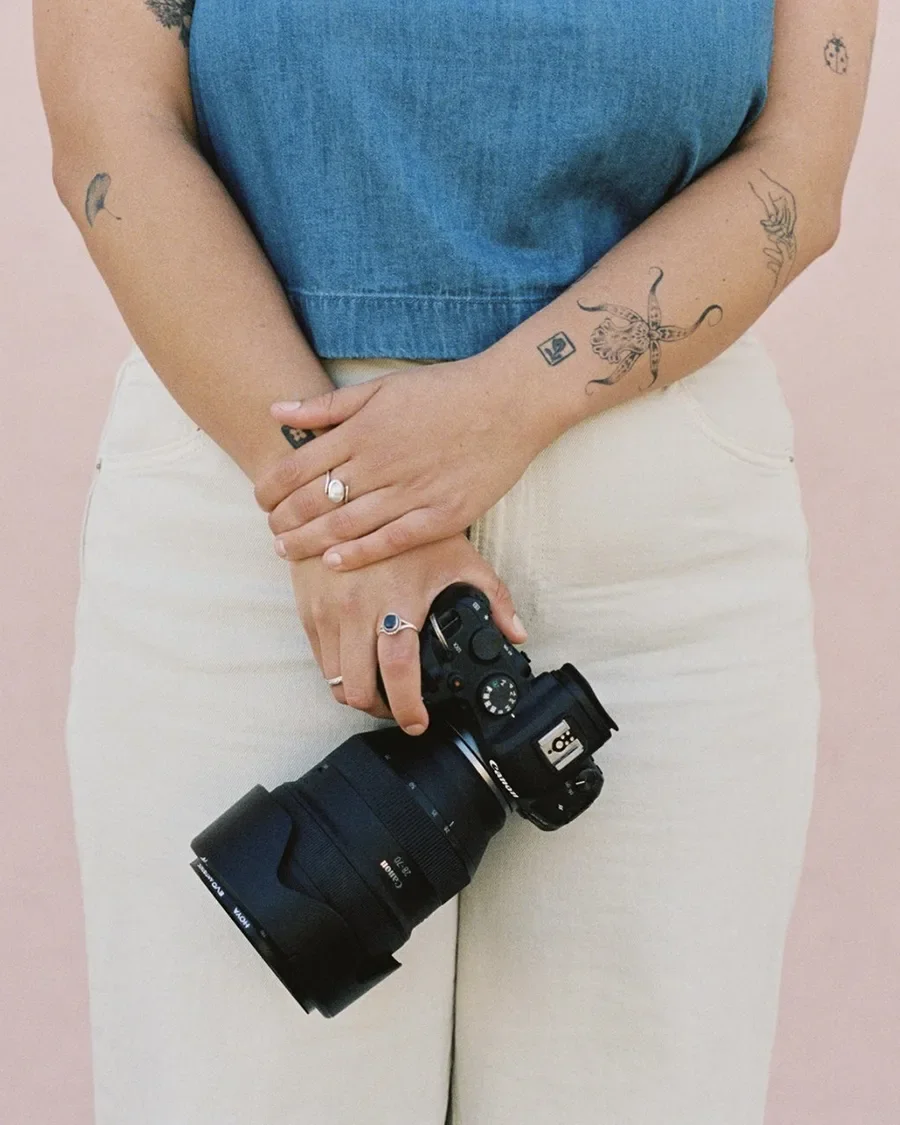 Zoe wearing a blue top and beige pants holding a camera with tattoos on her arms, standing against a light pink background.