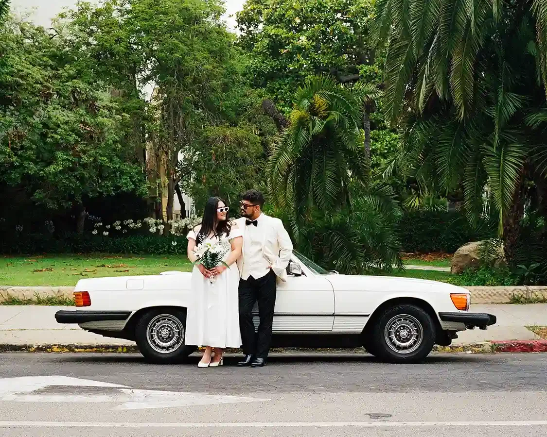 A couple dressed in formal attire standing beside a white vintage convertible car on a street with lush green trees in the background.