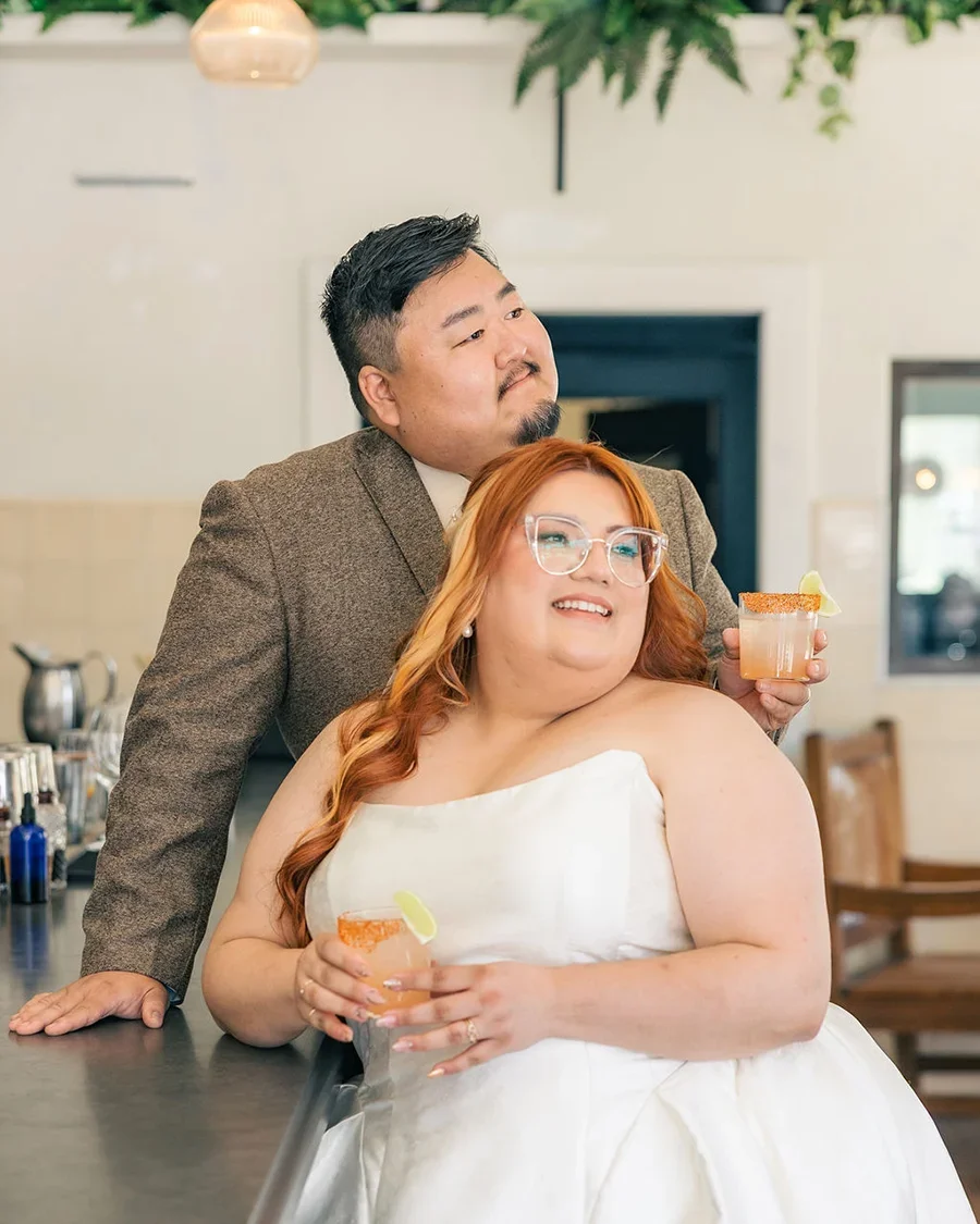 A couple, one with ginger hair and glasses and one with short black hair, is smiling and holding cocktails with lime wedges in a cozy bar or restaurant with hanging light fixtures and greenery.