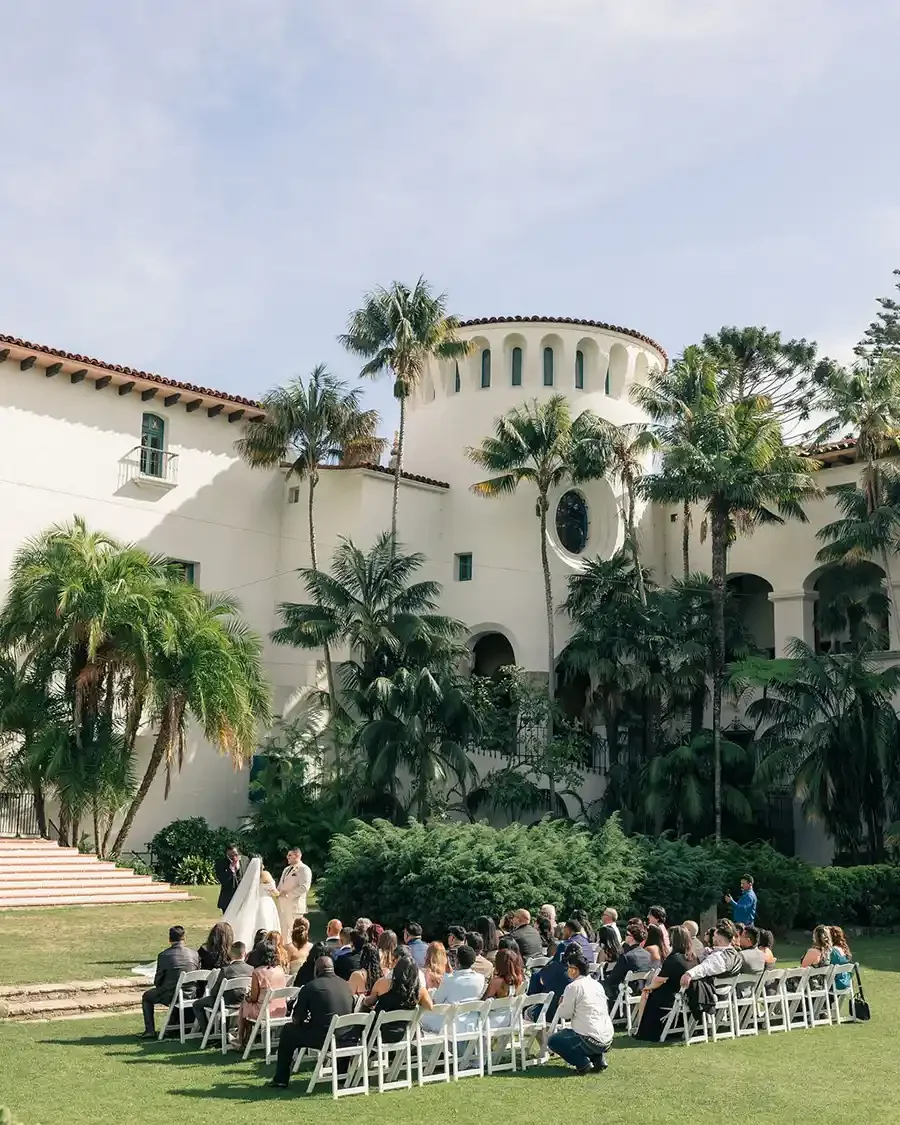 Outdoor wedding ceremony with guests seated on white chairs, couple standing in front of officiant, surrounded by palm trees and a large white building.