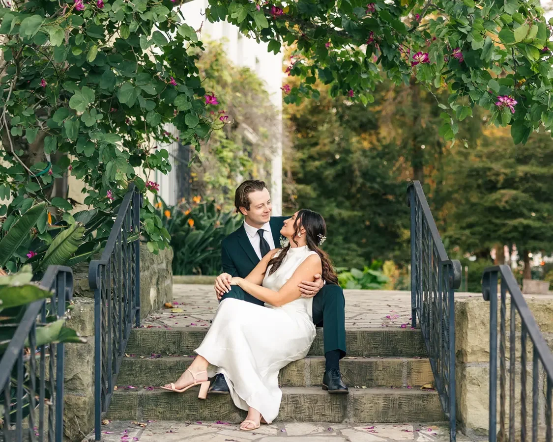 A couple sitting on stone steps in a garden, embracing and looking at each other, with trees and flowers around.