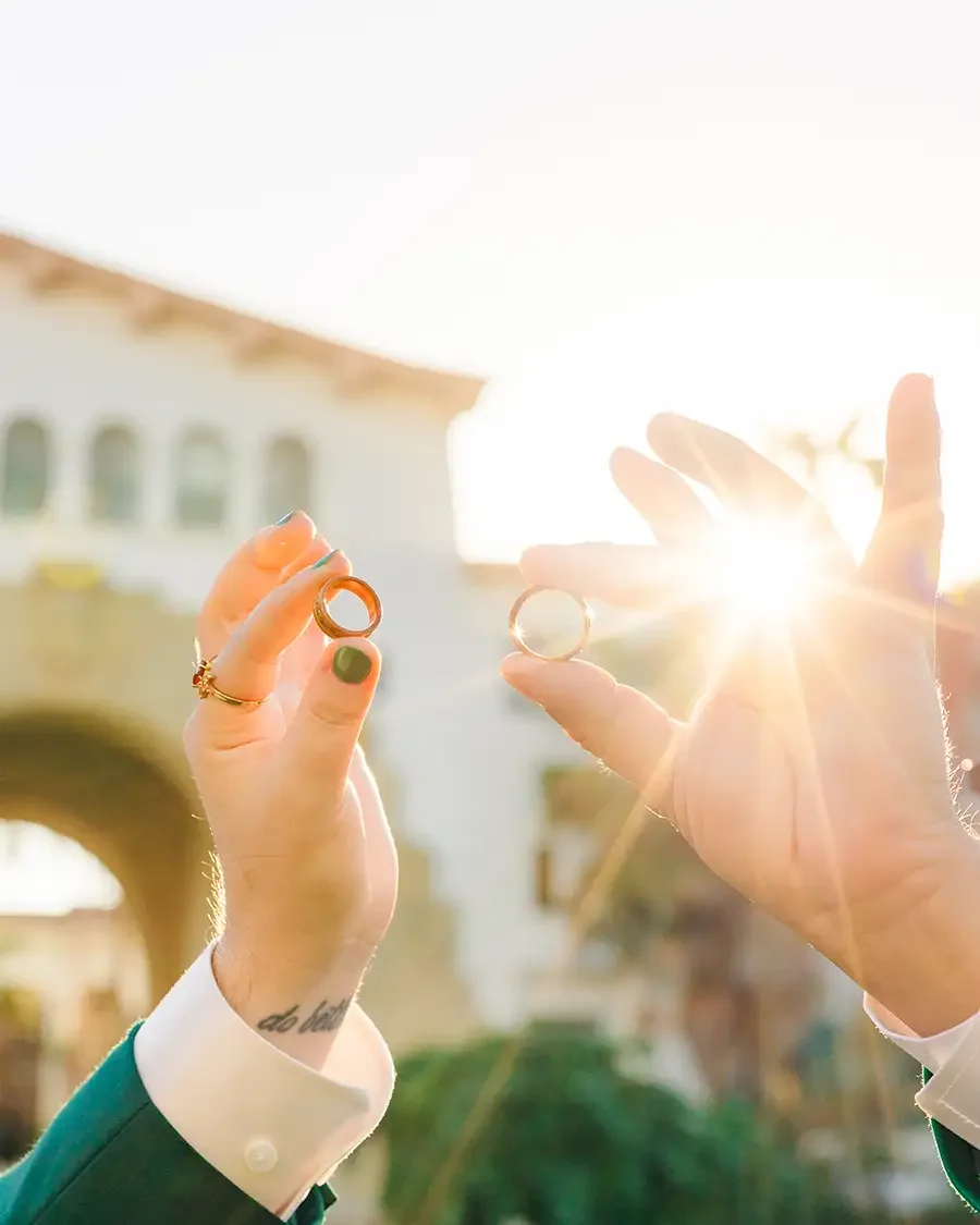 Two people holding up wedding rings outdoors at sunset, with one person's hand in focus and sunlight shining behind the rings.