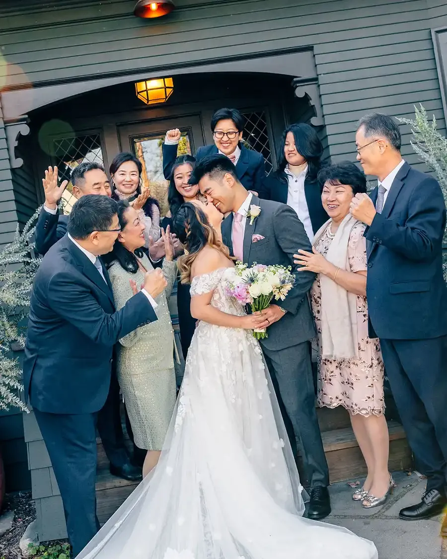 A group of people celebrating a wedding outside a house, with a bride and groom in the center. The bride is holding a bouquet and wearing a white wedding dress, while the groom is in a gray suit. The group is smiling, clapping, and cheering.