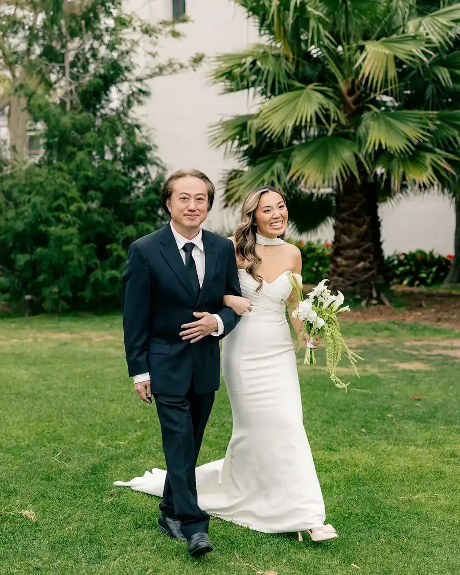 A bride in a white wedding gown walking arm-in-arm with a man in a navy suit on a lush green lawn with palm trees and other greenery in the background.