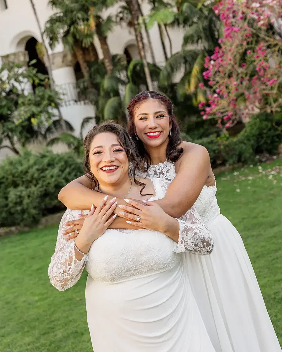 Two women dressed in white wedding gowns smiling and embracing outdoors in a garden setting with greenery and pink flowers.