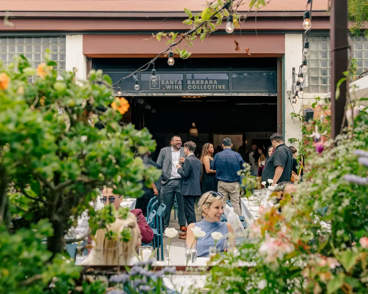 People gathered at an outdoor event near a building with a sign that says 'Santa Barbara Wine Collective.' Some are seated at tables; others are standing and chatting. There are string lights hanging above, and the scene is decorated with greenery and flowers.