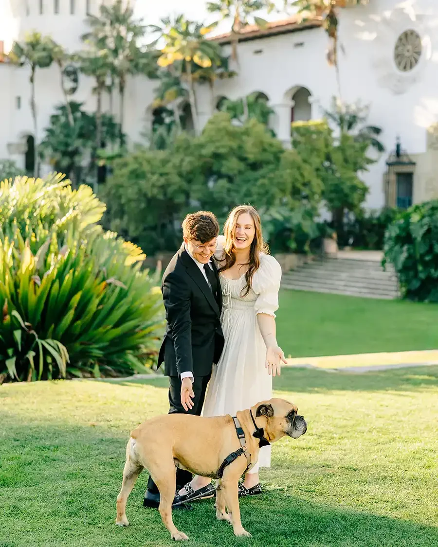 A couple dressed in formal attire, standing on a lawn with a large dog wearing a harness. They are smiling and appear to be enjoying themselves in a garden or park setting.
