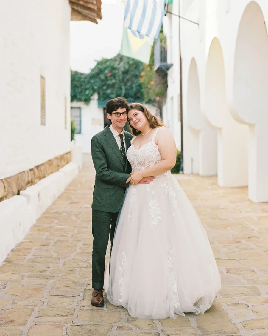A newlywed couple stands closely together in a narrow, cobblestone alleyway. The groom is wearing glasses, a dark green suit, and a tie, while the bride is in a white wedding dress with lace details. They are smiling and holding hands in front of a white wall with arches and greenery in the background.