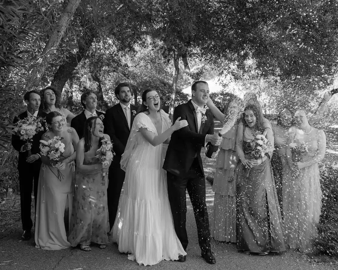 Black and white photo of a wedding celebration with the bride and groom opening champagne as friends and family laugh and cheer around them under trees.