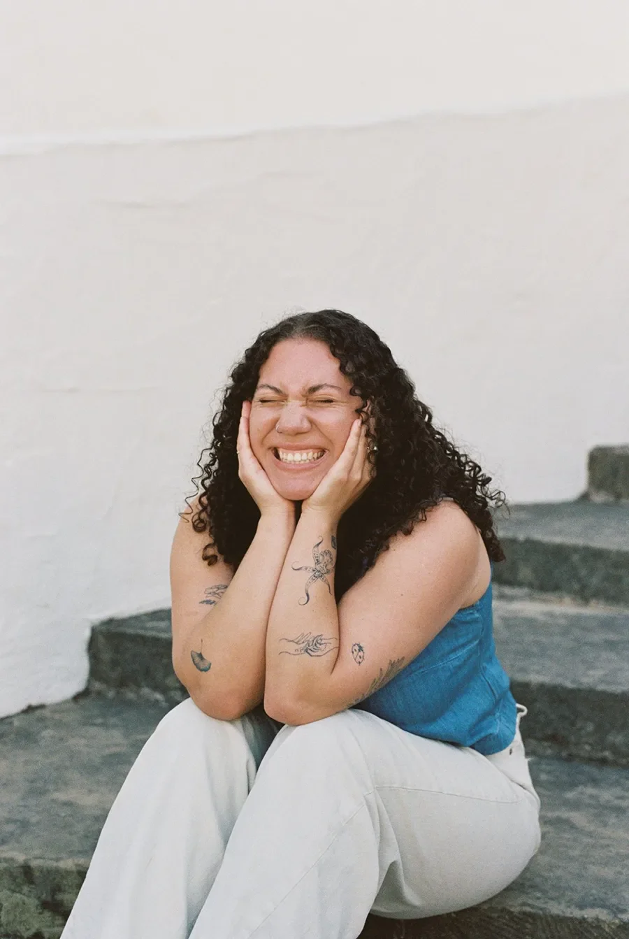 Photographer Zoe smiling sitting on concrete steps against a white wall, wearing a blue top and white pants.