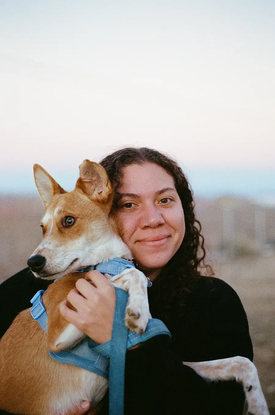 Photographer Zoe smiling and holding a tan and white dog wearing a blue harness outdoors at sunset.