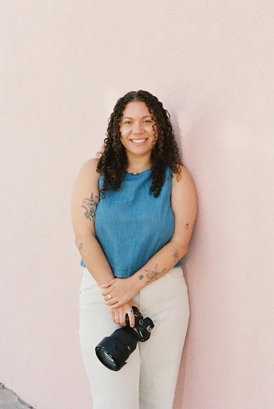 Zoe wearing a blue sleeveless top and white pants, standing against a pink wall, holding a camera with a large lens, smiling at the camera.