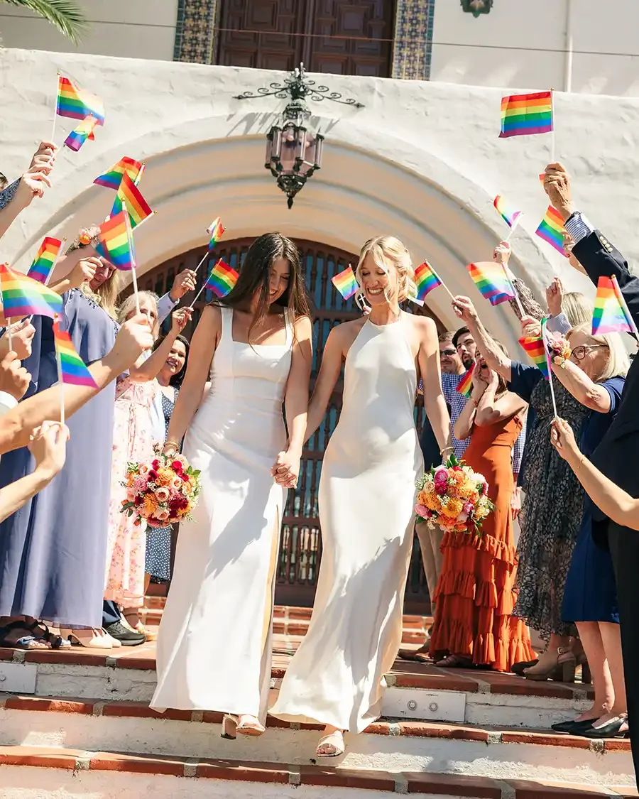 Two women in white dresses holding hands and bouquets, walking down the steps of a building, surrounded by people holding rainbow flags, celebrating a wedding or pride event.