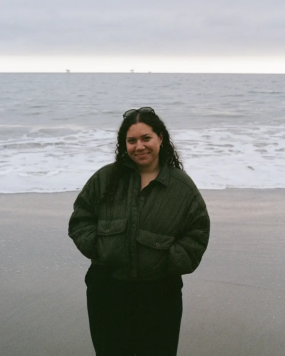 Zoe standing on a beach with ocean waves in the background, wearing a dark jacket and sunglasses on her head.