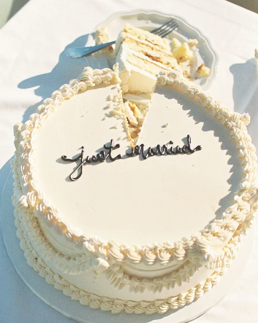 A white heart-shaped cake with a border of whipped cream, decorated with the words "Just Married" in black cursive icing. A slice is cut out, revealing layers of cream and filling. Behind the cake, there is a plate with a partially eaten slice of cake and a fork resting on it.