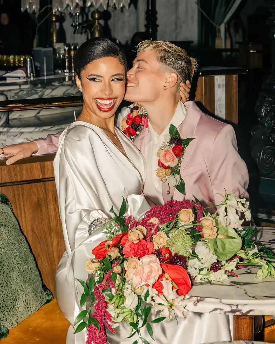 A happy couple at their wedding celebration. The bride is smiling and wearing a white satin dress, while the groom kisses her on the cheek. They are sitting at a table decorated with a large bouquet of pink, red, peach, and white flowers. The background suggests an elegant indoor setting with dim lighting.