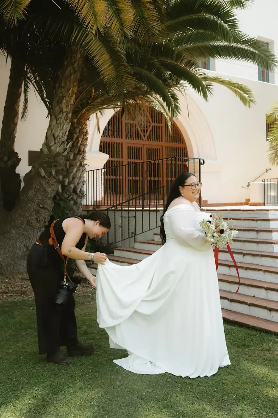 A bride in a white wedding dress holding a bouquet of flowers, smiling, as photographer Zoe adjusts her dress under a tall palm tree outside a building with stairs and an arched wooden door.