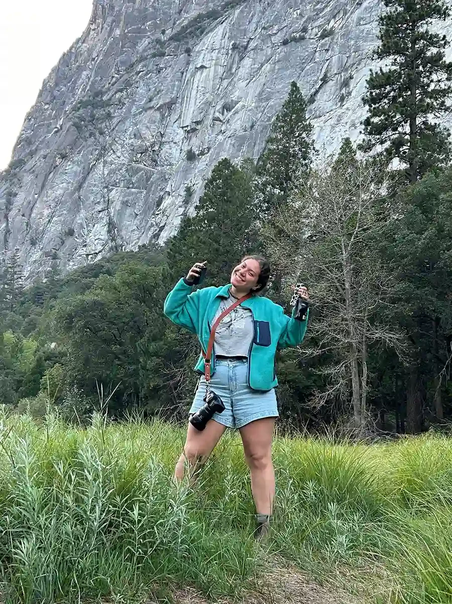 Zoe standing in a grassy area in front of a forest with a rocky cliff in the background, smiling, holding a camera in one hand and a can in the other, wearing a teal jacket, jean shorts, and boots.