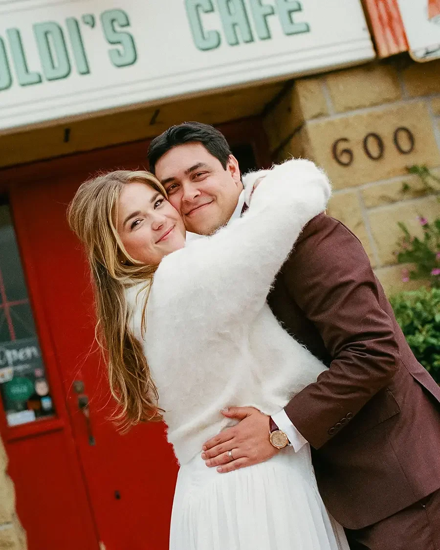 A man and woman hugging in front of a building with a sign that reads "GOLDI'S CAFE" and the number 600. The woman has long blonde hair and is wearing a white sweater and skirt. The man has dark hair and is wearing a brown suit with a white shirt. He is also wearing a watch.