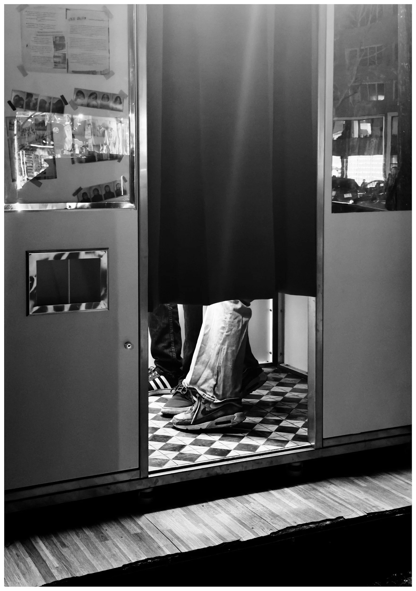 A person sitting on a stool at a booth, with only their legs and shoes visible, inside a small space with checkered flooring and reflective glass doors.