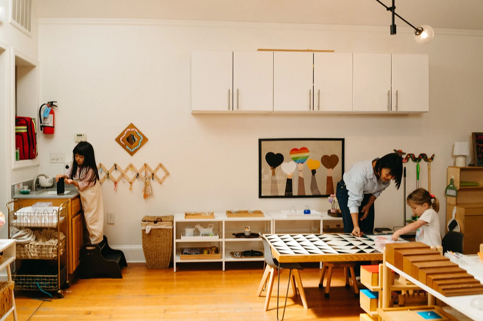 Montessori teacher leaning in to guide a preschooler at a tiled work table, with practical life shelves, a framed print of raised hands holding colorful hearts, and the brown stair sensorial material in the foreground at Little Eden in San Francisco