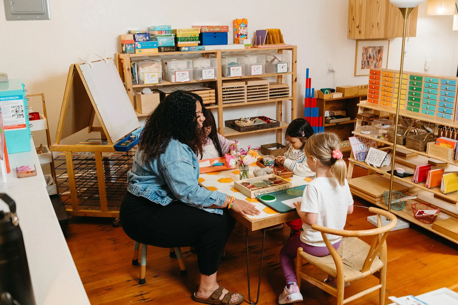 Montessori teacher and preschoolers threading beads together at a table filled with flowers and art materials