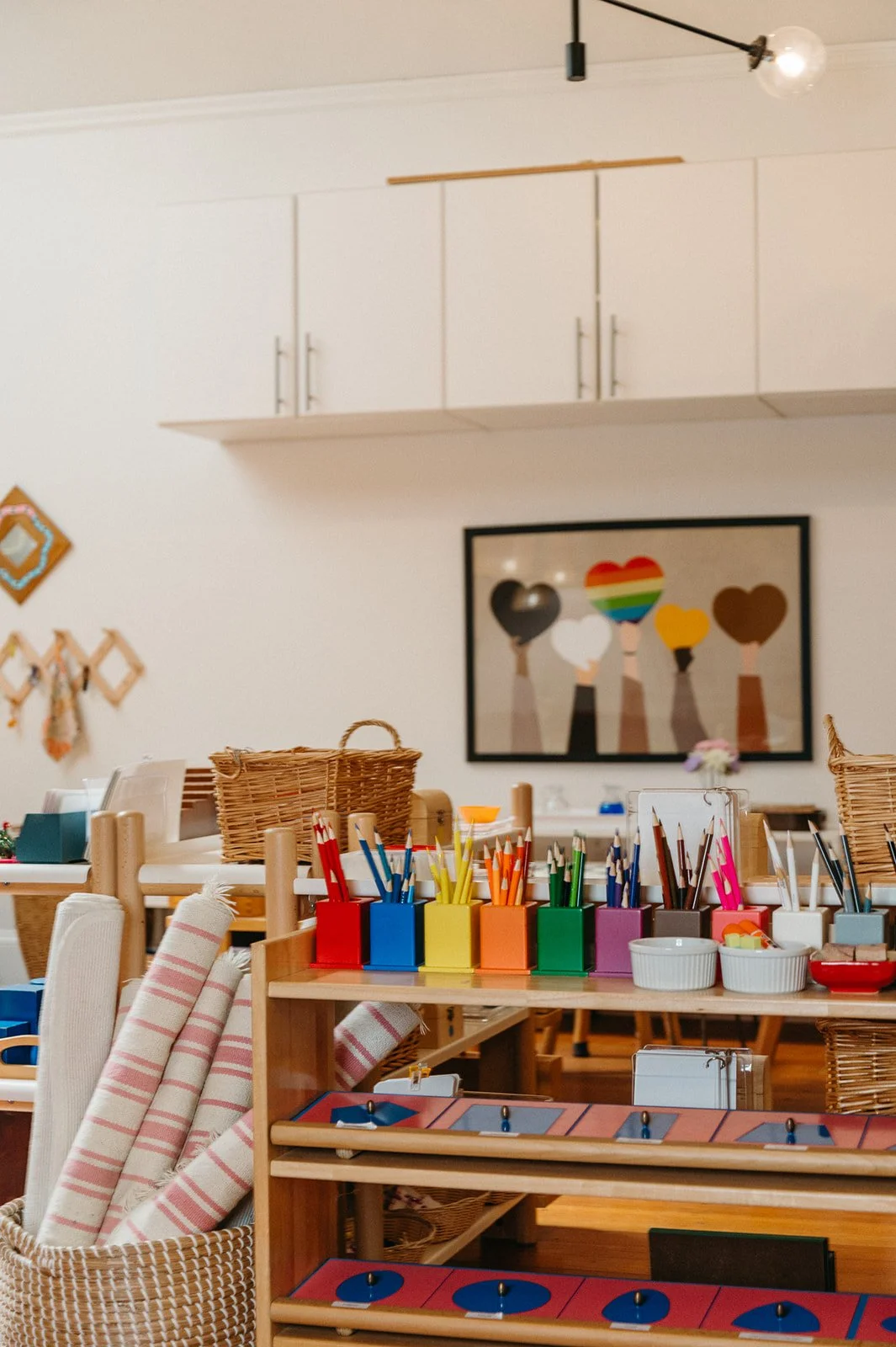 Colored pencils sorted into matching color-coded cups on a classroom shelf at Little Eden's arts-focused Montessori preschool in San Francisco, with woven baskets and a framed hearts print in the background