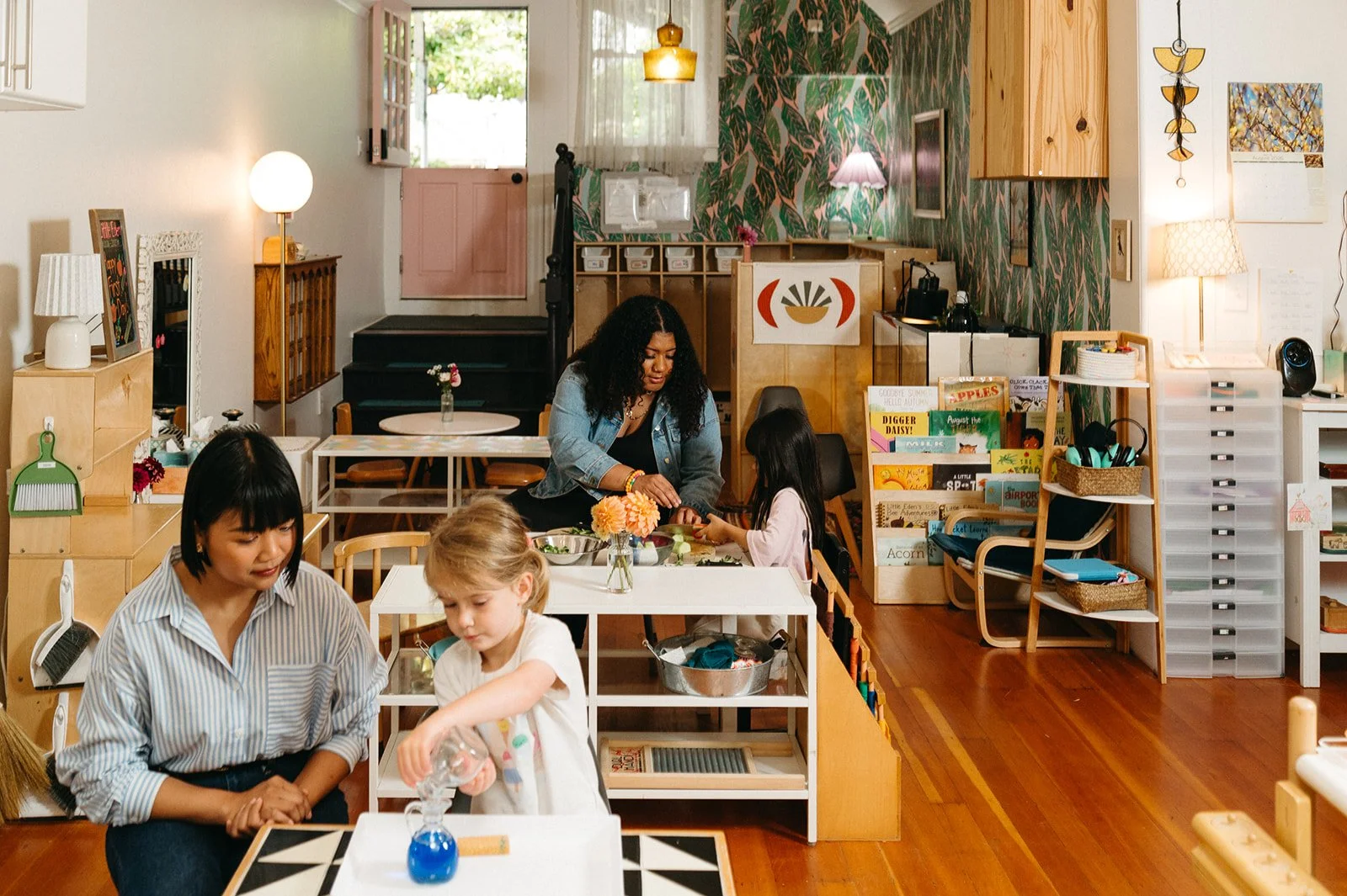 Montessori classroom at Little Eden in San Francisco, where a teacher watches a preschooler pour water between small glass pitchers while another teacher and child prepare food together, with a bookshelf of children's picture books nearby