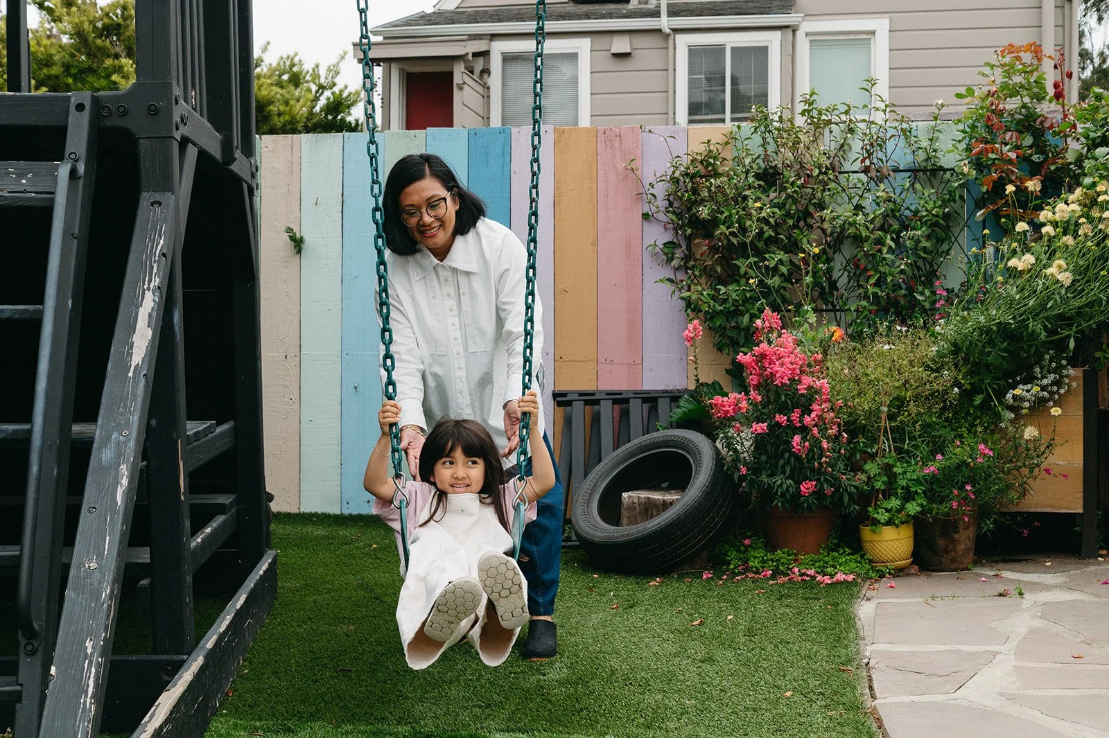 Little Eden Director, Michelle, pushing a smiling child on a swing in Little Eden's outdoor play space, with a colorful fence and flowers