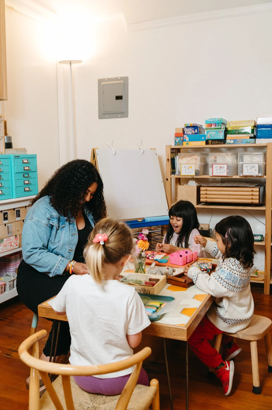 Montessori teacher and three preschoolers threading beads together at Little Eden in San Francisco, with flowers and art materials on the table