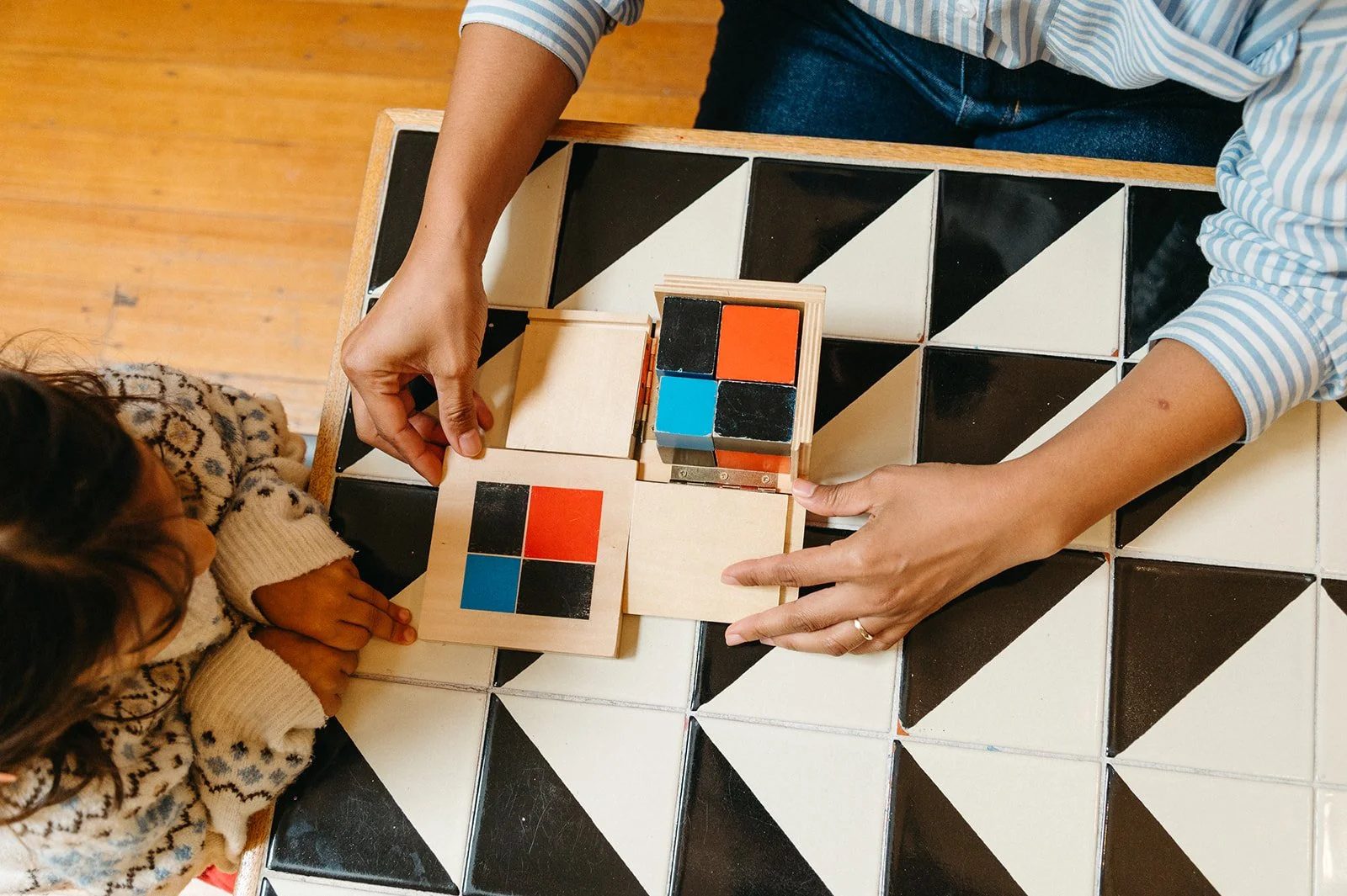 Overhead view of a Montessori teacher presenting the binomial cube to a preschooler, opening the wooden box to reveal red, blue, and black blocks at Little Eden Montessori preschool in San Francisco