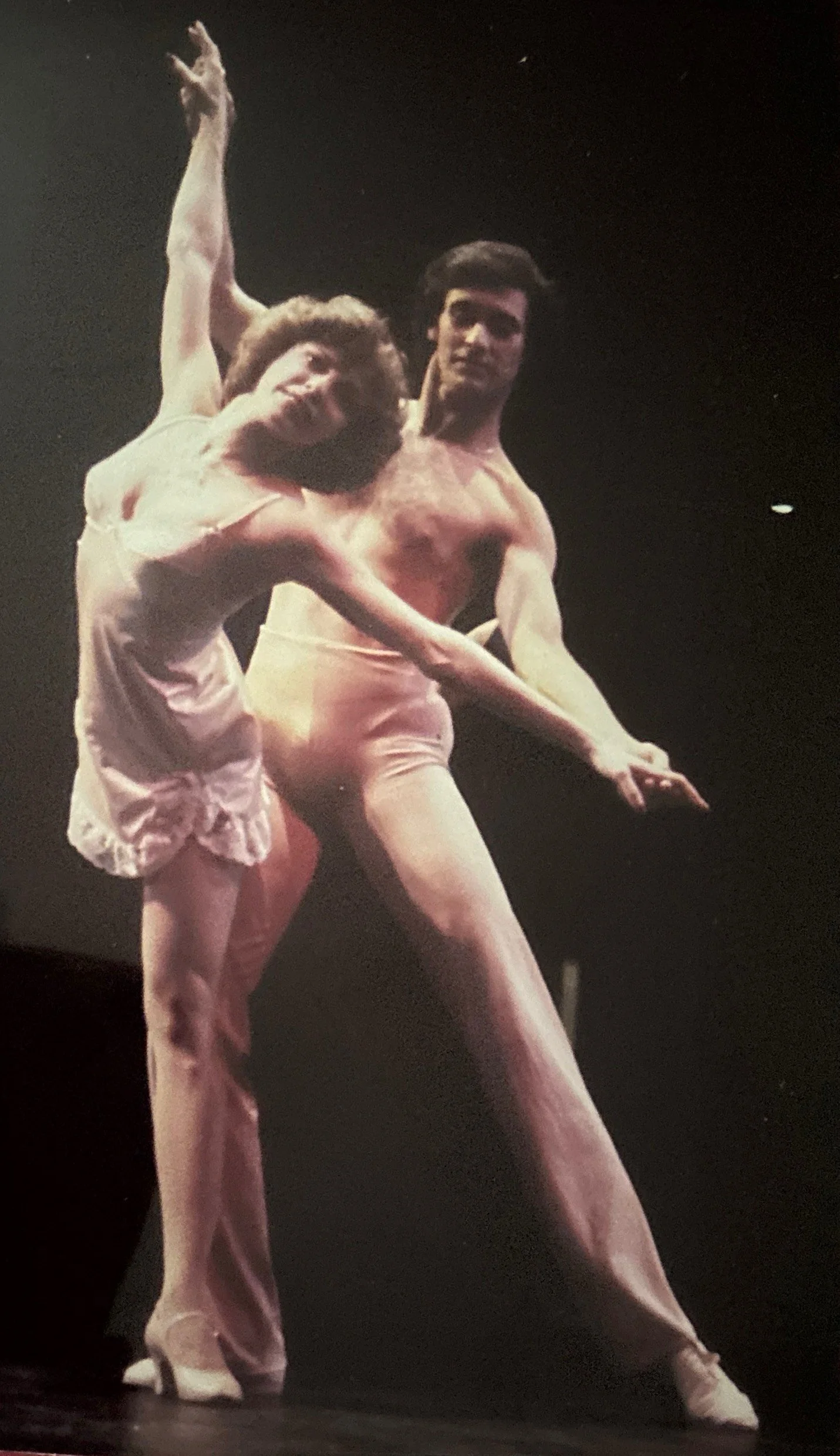 A ballet dancer lifts a female ballet dancer in a dance pose on a dark stage.