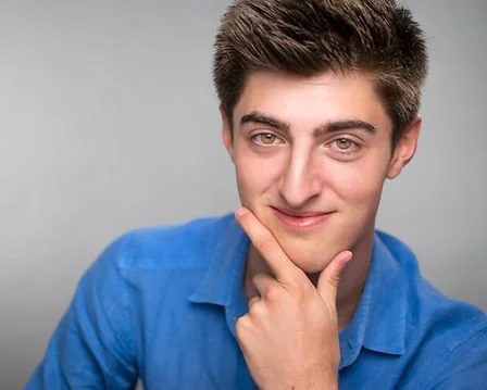 Young man with short brown hair, wearing a blue shirt, posing with his hand on his chin and smiling.