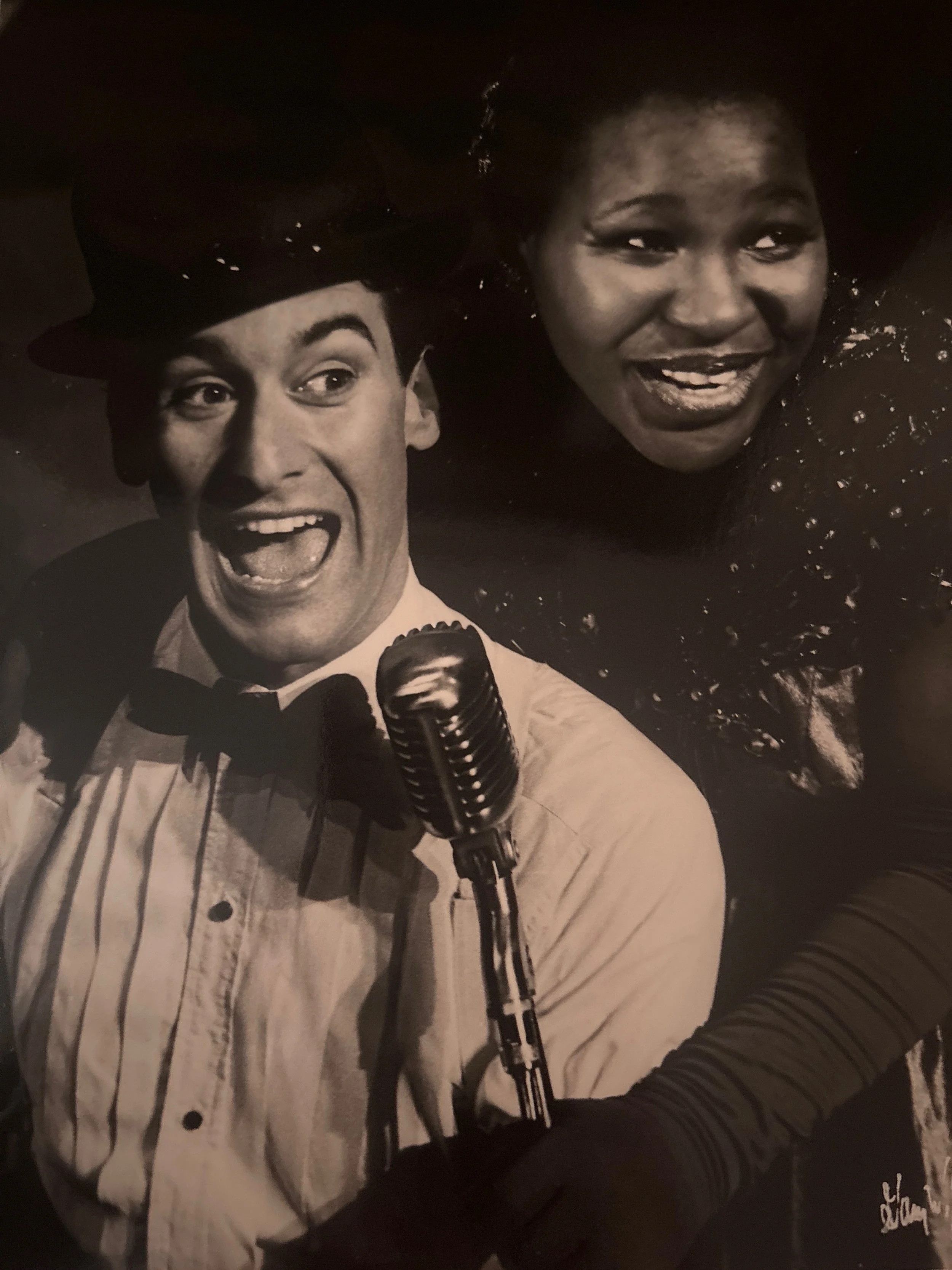 Black and white photo of two women smiling and looking at each other, one holding a vintage microphone.