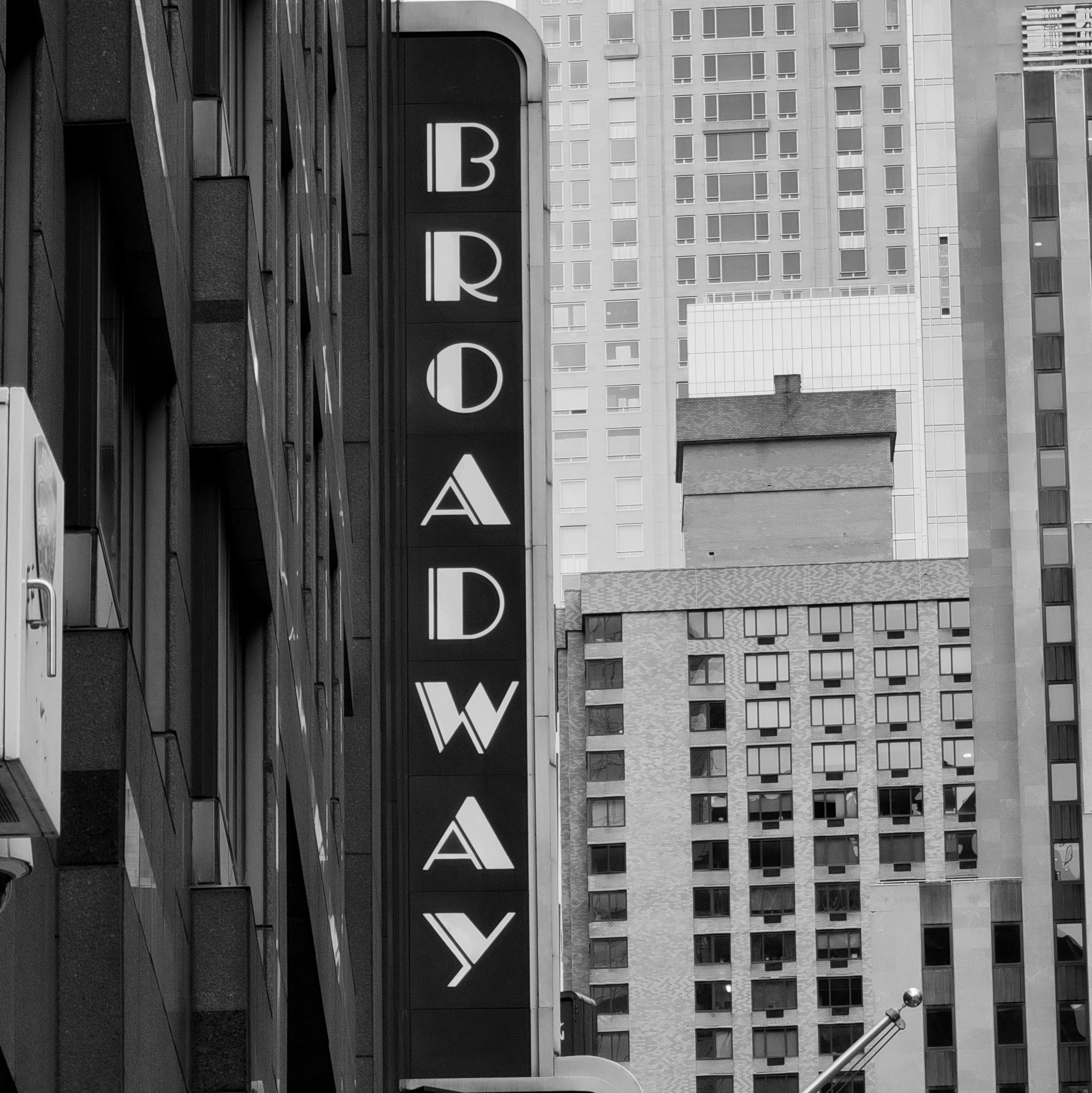 Black and white photograph of a cityscape showing a building with a vertical sign displaying the words "BARD" and a background of high-rise buildings with numerous rectangular windows.