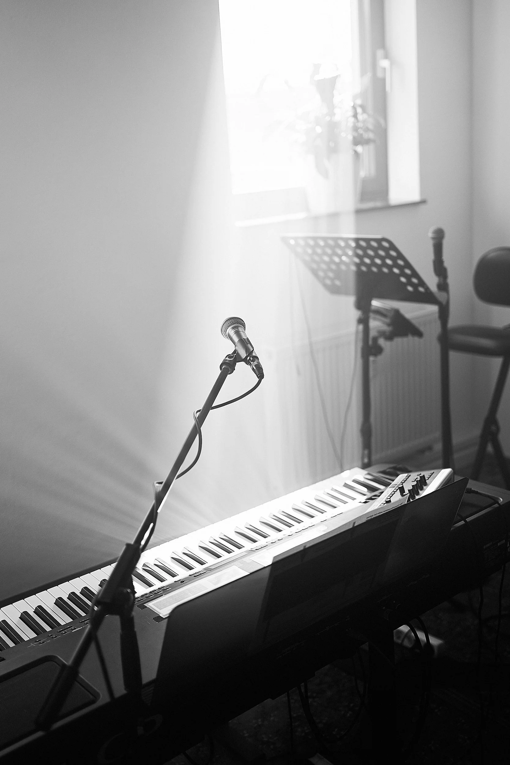 An electronic keyboard on a stand with a music sheet, microphone, and pop filter, set up in a room with a window and a chair in the background.