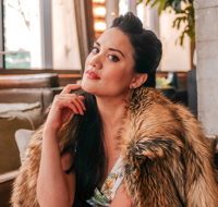 Woman with dark hair wearing a fur coat, sitting indoors with a window and plants in the background.